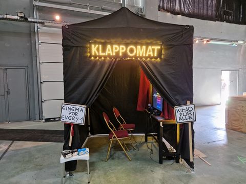 The photo shows a so-called Klappomat, a small tent in which two chairs and a screen with headphones have been set up. The installation allows one or two people to get to know an audio description for the visually impaired or subtitles for the hearing impaired and guides them through the process step by step. The users learn how difficult it is to describe a complex scene in short words and what requirements successful film descriptions have to meet.