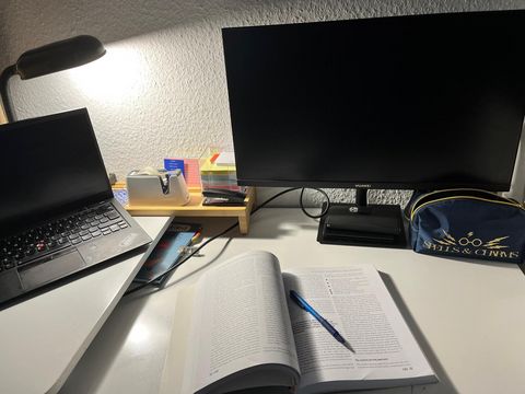 A desk with a laptop and desktop, a book lies open in front of it. It's dark, the desk lamp is on.