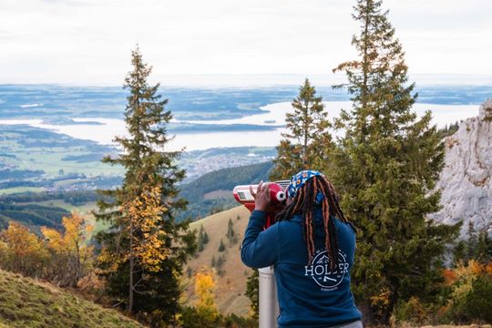 Doris steht in der Natur und auf einer Anhöhe. Sie schaut durch ein Fernglas auf den Chiemsee vor ihr.