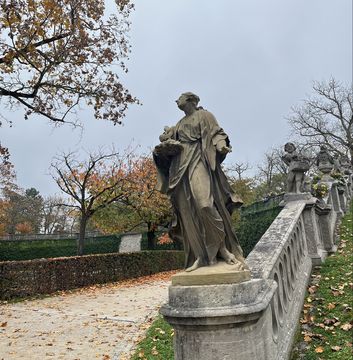 You can see the park of the Würzburg Residence. The weather is grey and foggy. In the foreground you can see a statue, the paths are covered with autumn leaves.