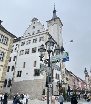 You can see the pedestrian zone of Würzburg's old town. Lots of people are walking around there, the sky is grey and cloudy. You can see lots of historical and pretty details, like street lamps and street signs, pretty historical buildings and towers.