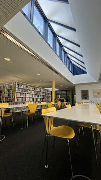 In the foreground you can see large tables for reading and working, with yellow chairs. In the background you can see many bookshelves filled with lots of books.