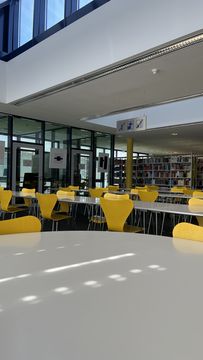 In the foreground you can see large tables for reading and working, with yellow chairs. In the background you can see many bookshelves filled with lots of books.