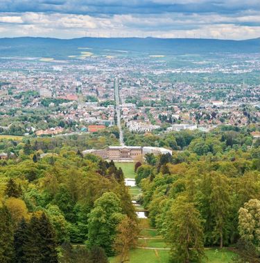 Blick auf das Schloss Wilhelmshöhe vor einem grünen Garten, im Hintergrund erstreckt sich die Stadt Kassel